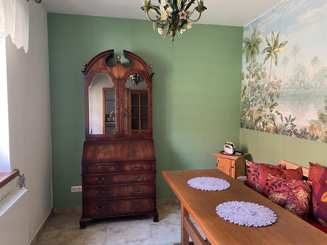 Ein Zimmer mit einem Holzkleiderschrank, einem Esstisch mit Untersetzern, einer Couch mit gemusterten Kissen und einem Wandgemälde einer tropischen Landschaft.