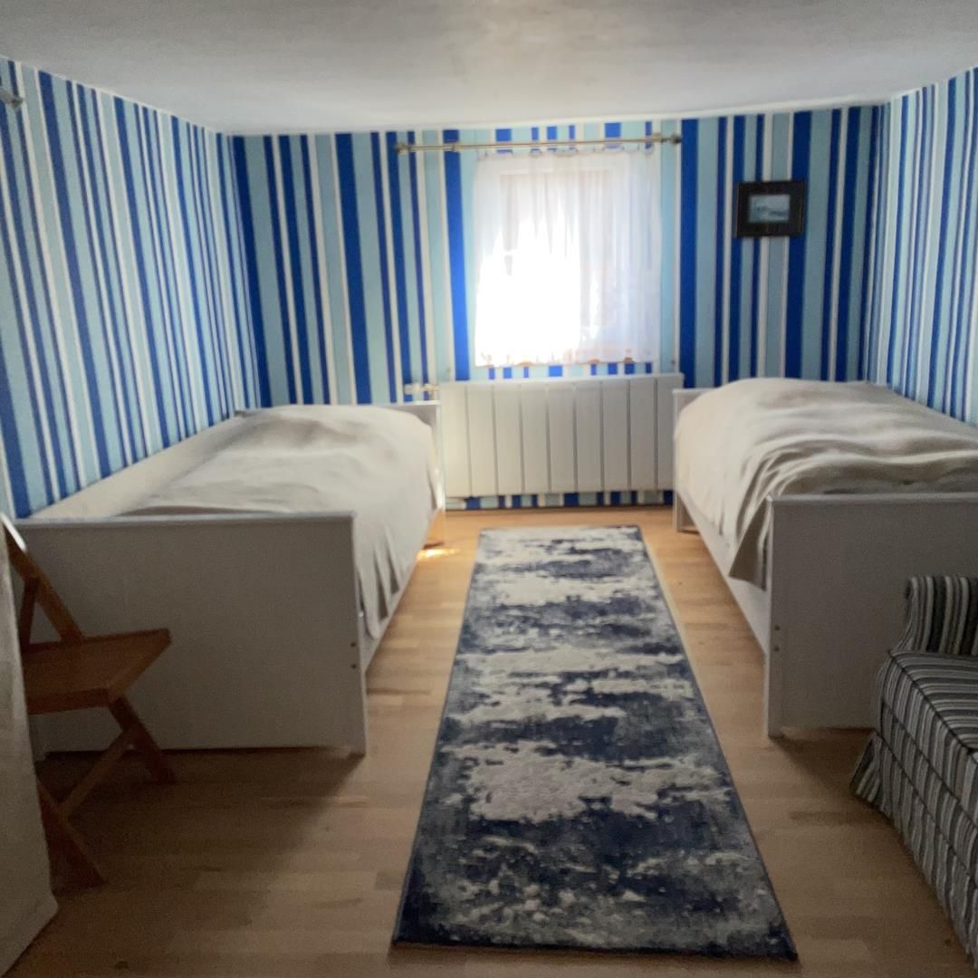 Ein Zimmer mit zwei Betten, einem Heizkörper, einem Fenster mit Vorhängen, einem Holzboden und einem blau gemusterten Teppich.
