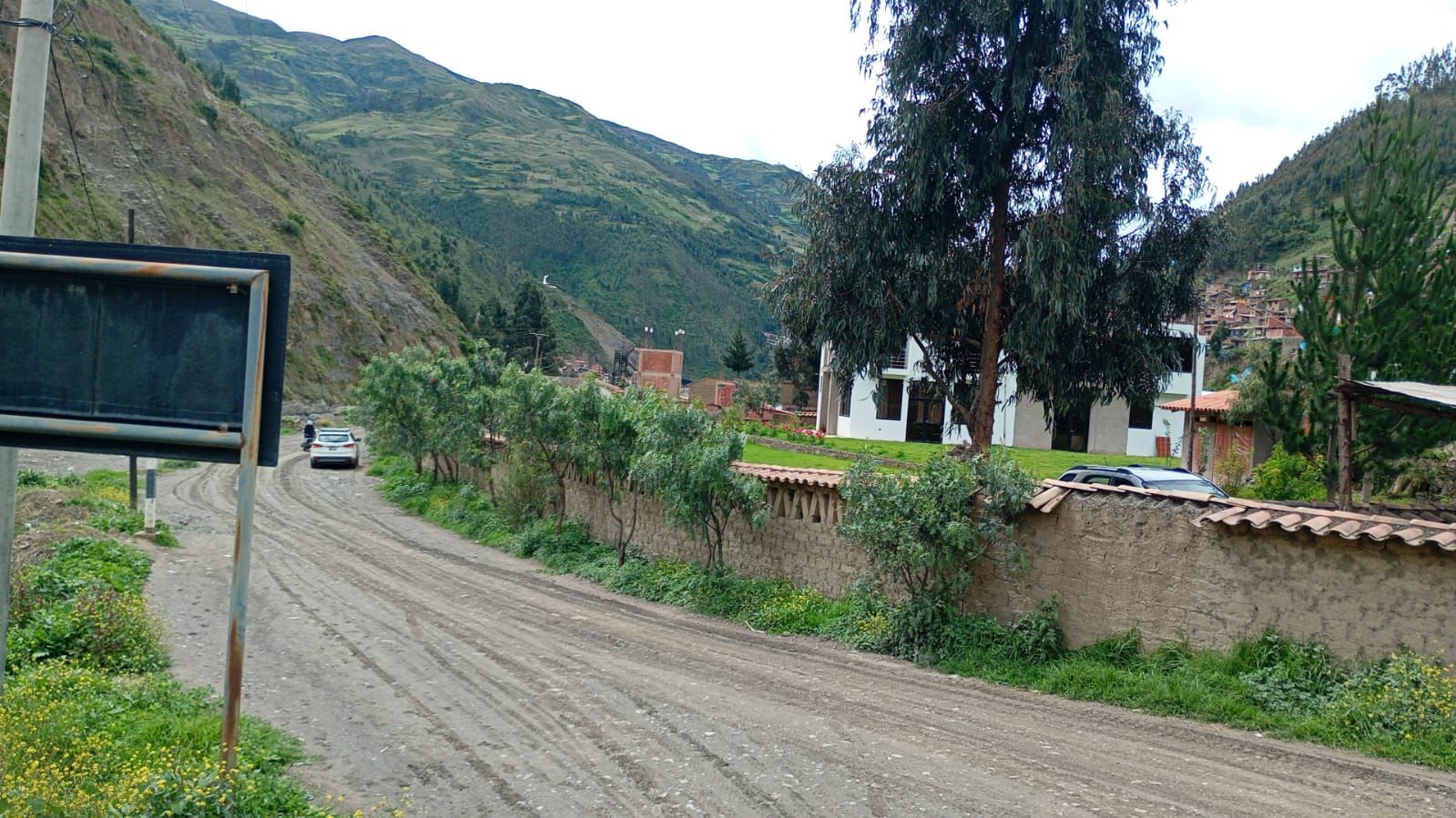 Un camino rural con un coche, casas con techos de tejas rojas y una montaña al fondo.
