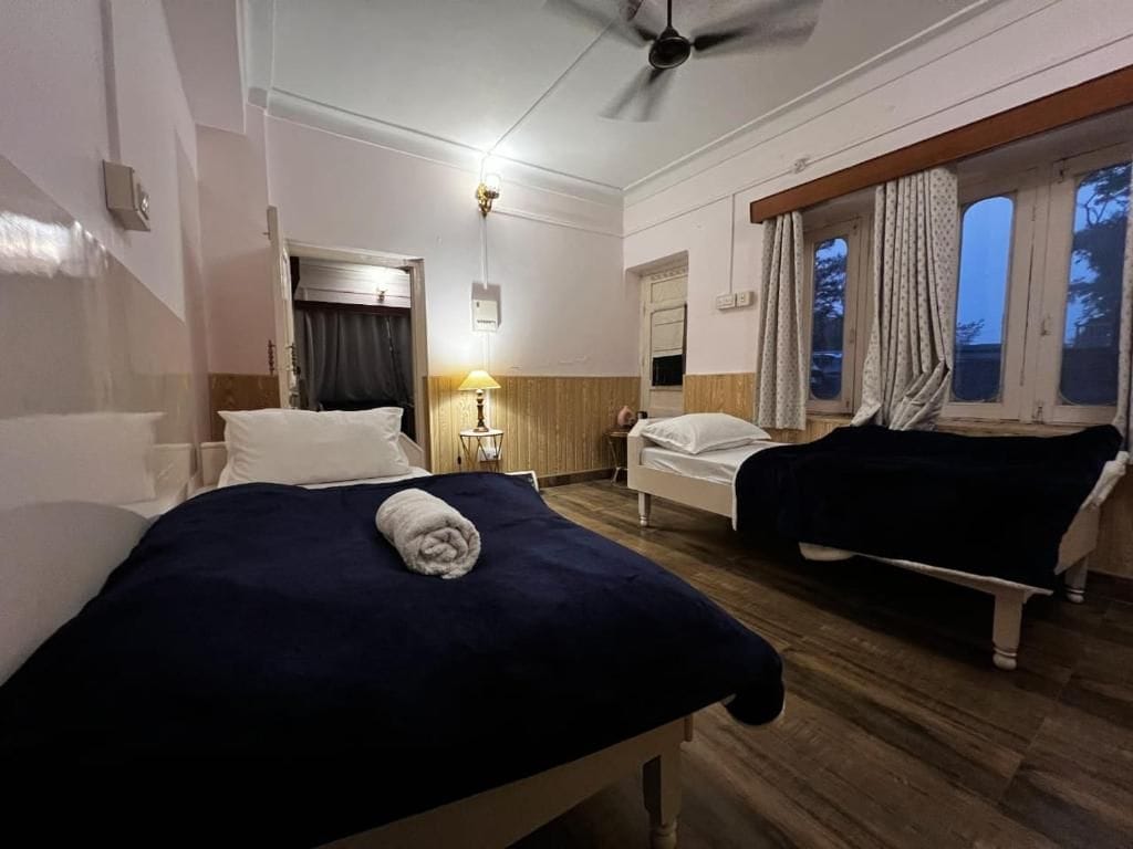 Una habitación de hotel con dos camas, un ventilador de techo, una ventana con cortinas y una mesita de noche con lámpara.