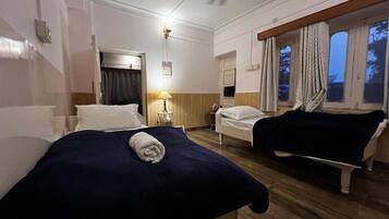 Una habitación de hotel con dos camas, un ventilador de techo, una ventana con cortinas y una mesita de noche con lámpara.