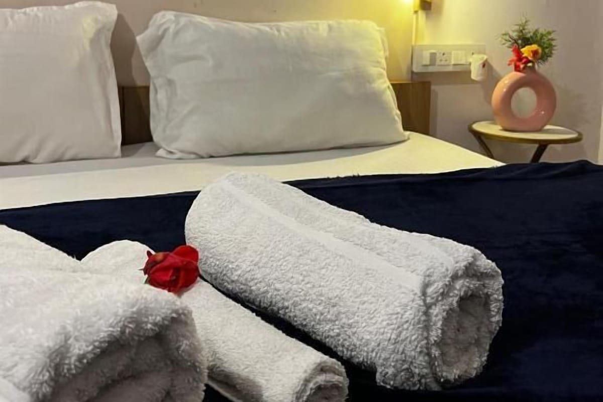 Una habitación de hotel con una cama, almohadas, una toalla enrollada con una rosa, un jarrón con flores y un aplique de pared.