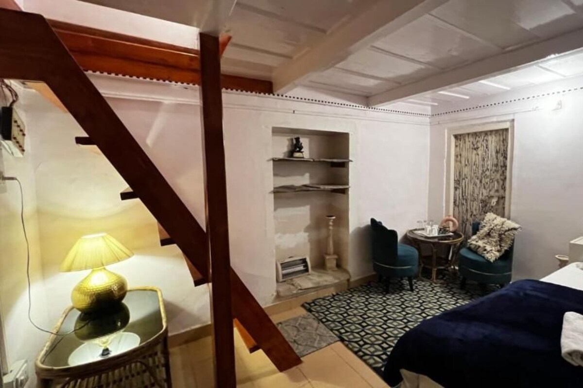 A hotel room with a bed, a chair, a small table with a lamp, a wardrobe, and a window with curtains.