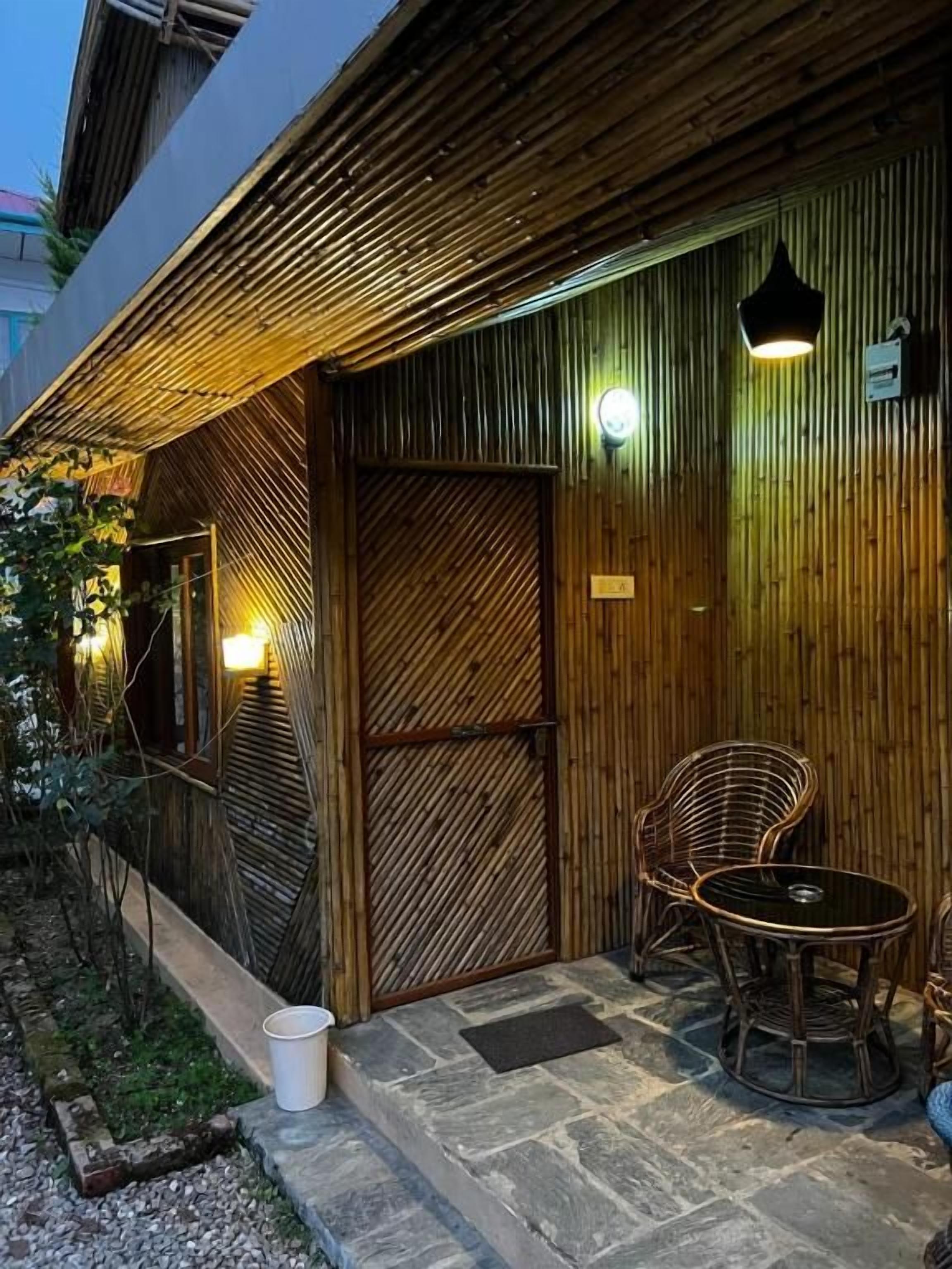Fachada de madeira com parede de bambu, porta de vidro e área de estar com cadeiras de vime e uma mesa.