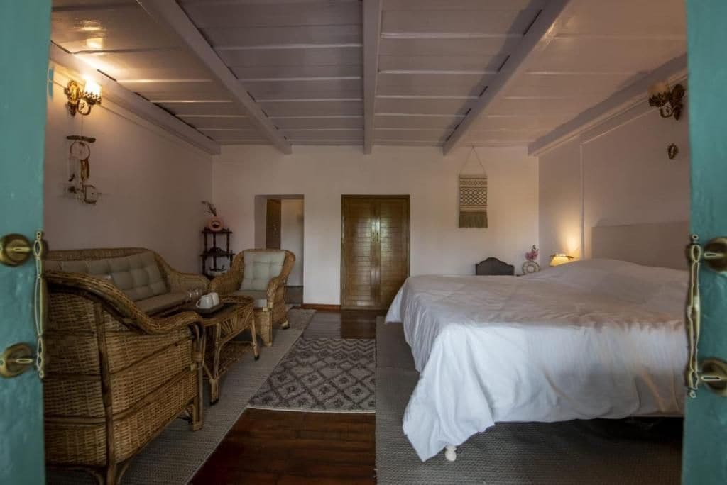 Ein Schlafzimmer mit einem Bett, einer Rattan-Couch, einem Sessel und einem kleinen Tisch. Eine Tür und eine wandmontierte Leuchte sind ebenfalls vorhanden.