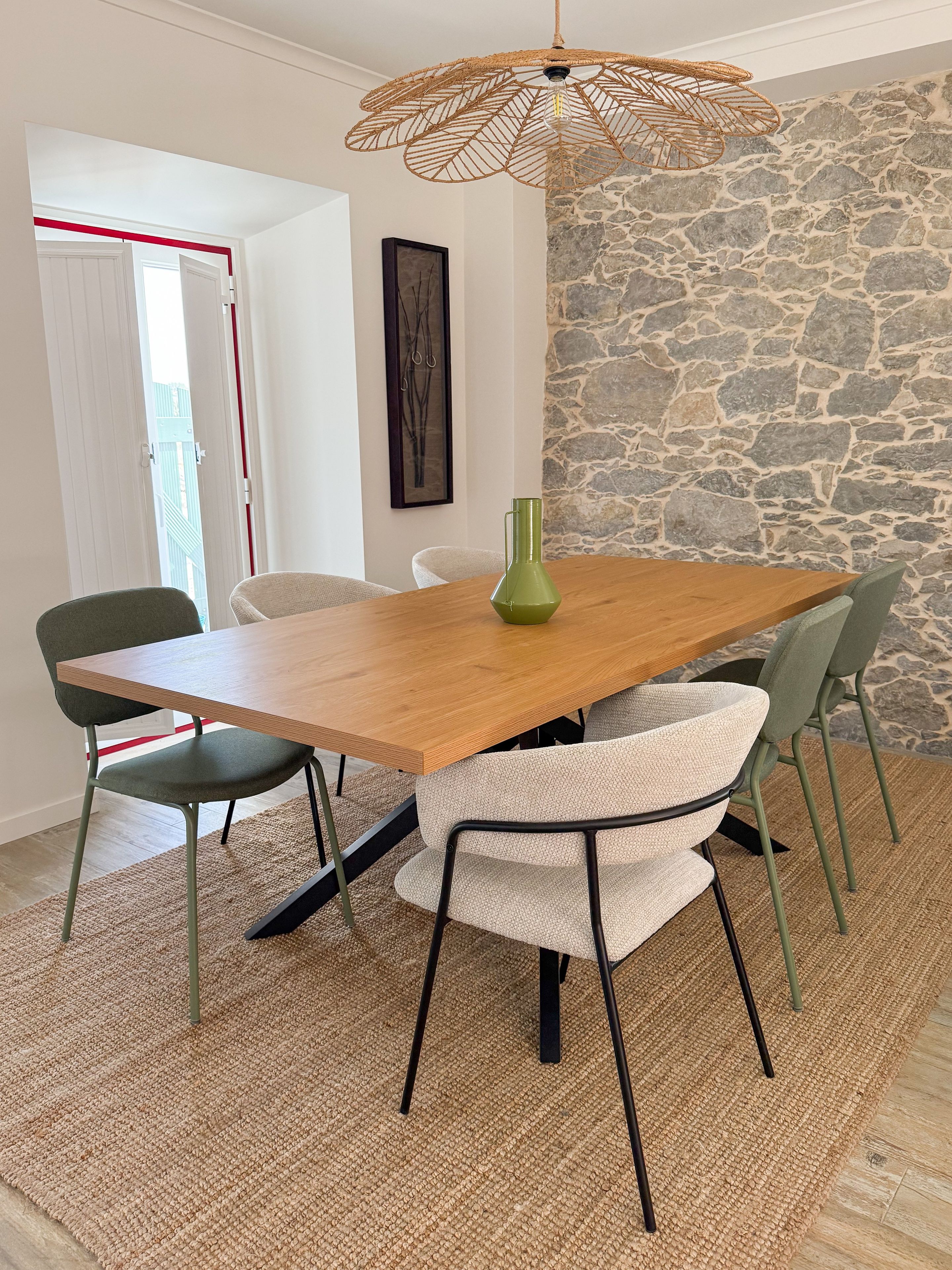 A dining area with a wooden table, four chairs, a woven pendant light, and a stone wall.
