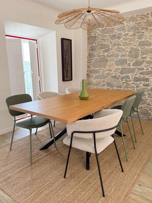 Un coin repas avec une table en bois, quatre chaises, un luminaire suspendu en fibres tressées et un mur en pierre.