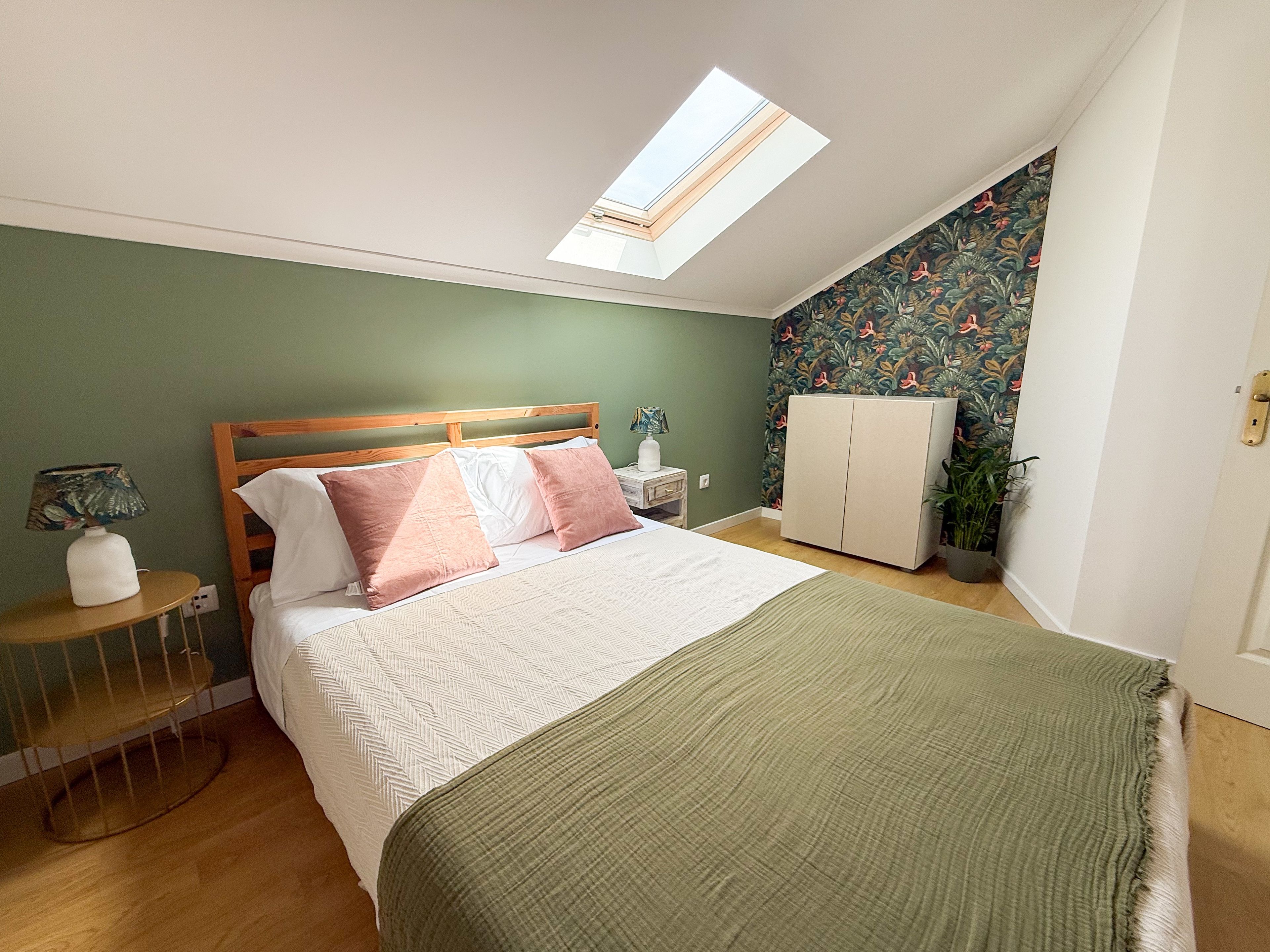Quarto com cama, claraboia, guarda-roupa, mesa de cabeceira, abajur e planta em vaso.