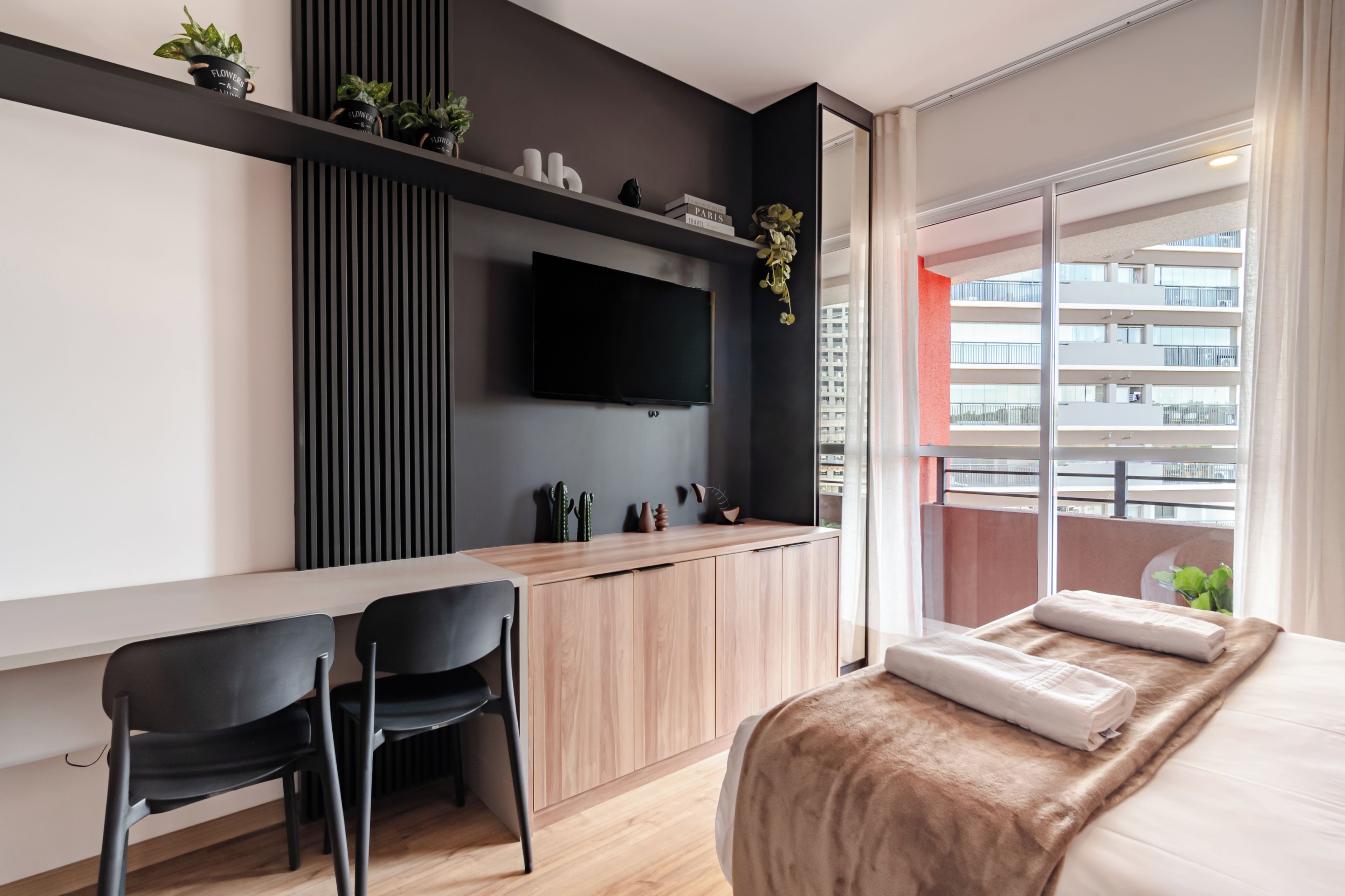 A modern bedroom with a bed, a desk with a chair, a TV, and a balcony with a view of the city.