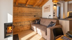 A cozy room with a wooden wall, a sofa with decorative pillows, a fireplace, and a kitchen area with a countertop. - Casa Quaranta - Two Bedroom Apartment, Sleeps 4 (Ponte di legno)