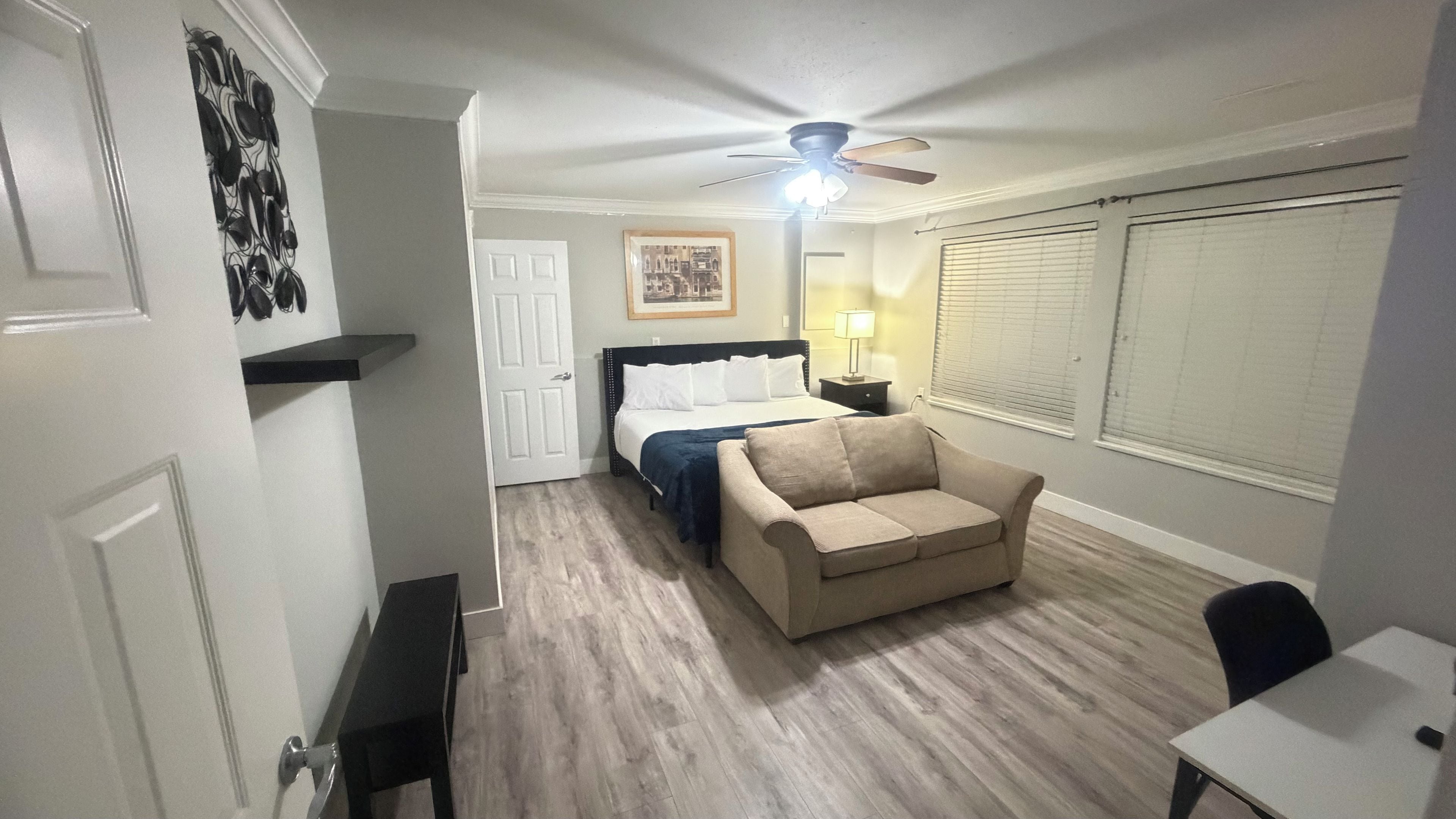 A hotel room with a bed, a sofa, a ceiling fan, a nightstand, and a window with blinds.