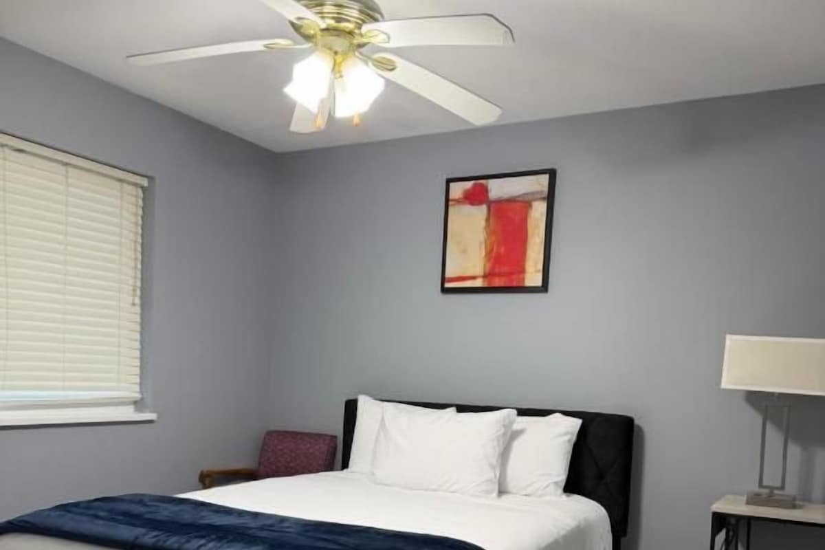 A bedroom with a bed, a ceiling fan, a wall-mounted lamp, and a window with blinds.