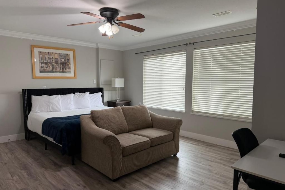 A bedroom with a bed, a sofa, a ceiling fan, and large windows with blinds.
