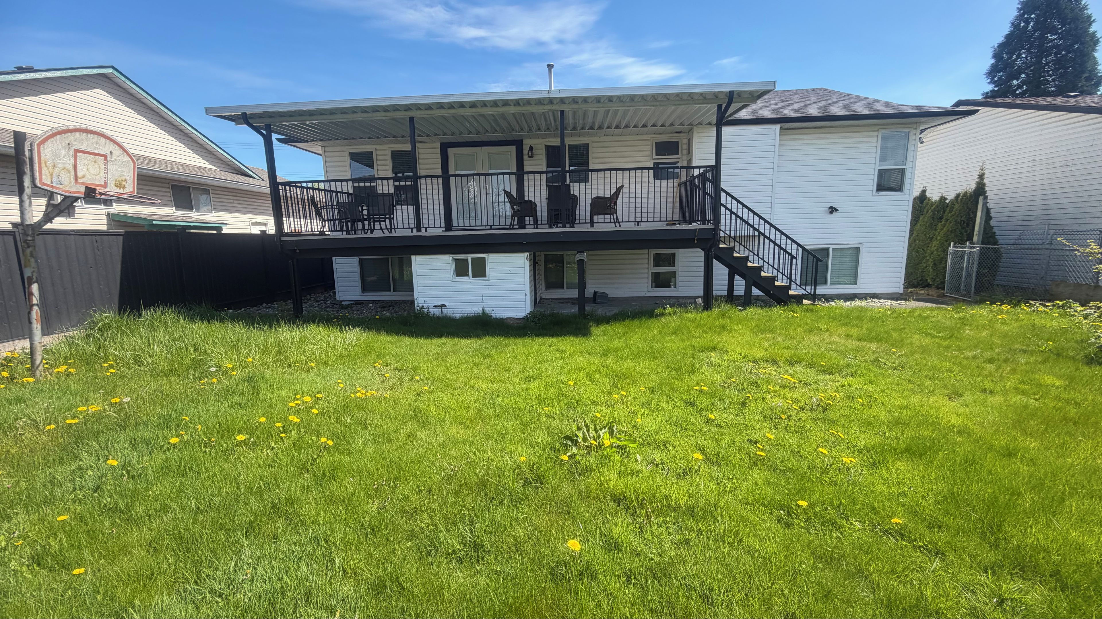 A two-story house with a balcony, a basketball hoop, and a grassy yard with dandelions.