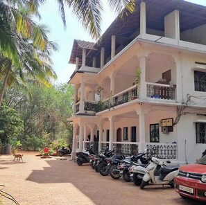 A multi-story building with a covered balcony area, a row of parked scooters, and a dog walking on the pavement. - Room-peaceful Stay Private Studio R-11 1 (Arambol)