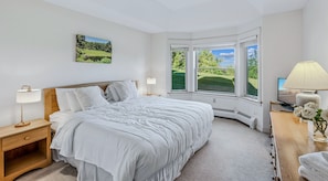 A bedroom with a large bed, two bedside tables with lamps, a desk with a lamp, a view of greenery through the windows, and a painting on the wall. - Solitude Ski-In/Ski-Out Condo with Pool & Hot Tub (Ludlow)