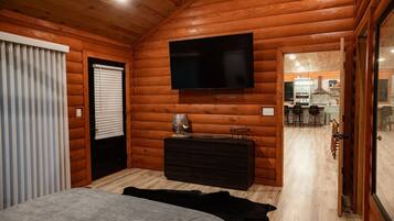 A log cabin bedroom with a bed, a dresser, a flat-screen TV, and a view into the kitchen area.