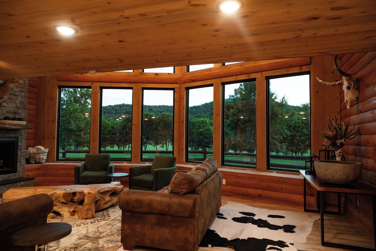 A log cabin interior with a fireplace, large windows, a sofa, and a cowhide rug.