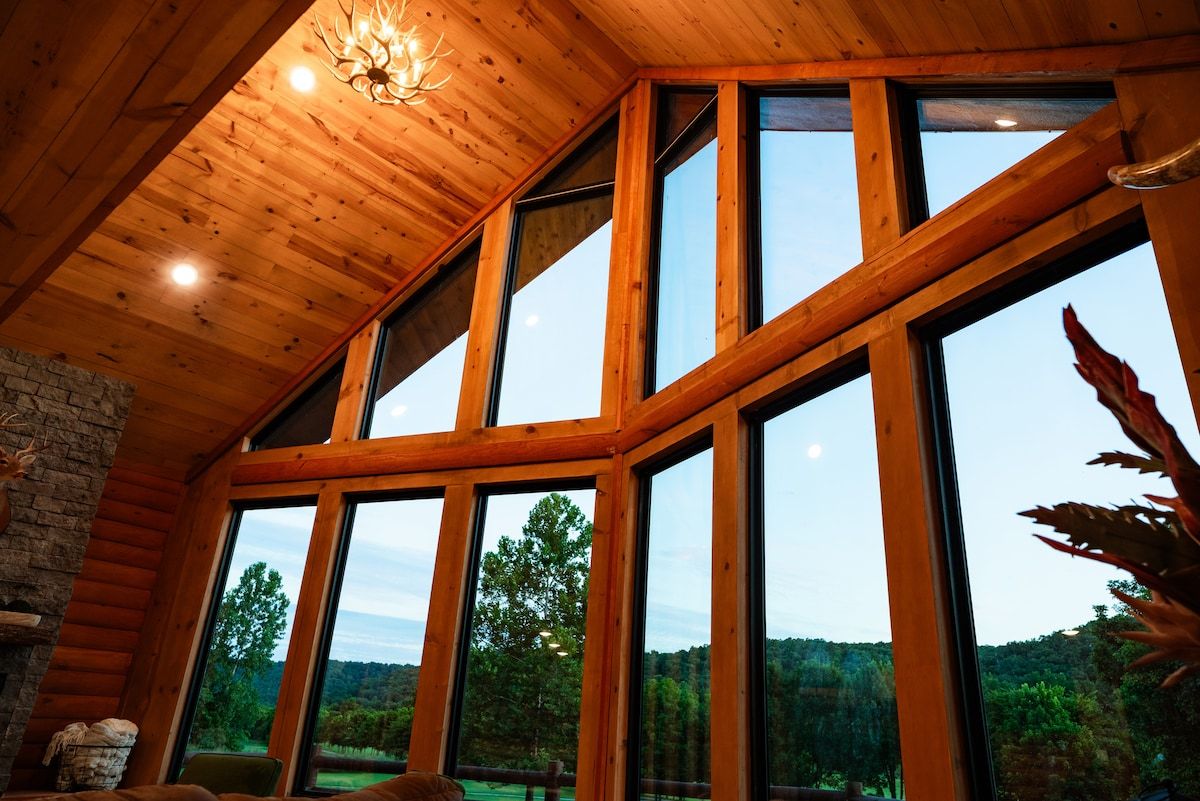 A cabin interior with large windows, wooden beams, and a stone wall.