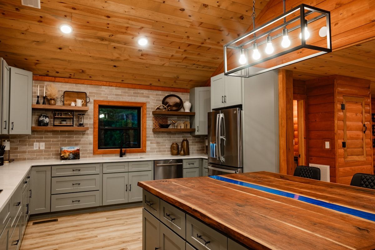 A modern kitchen with wooden ceiling, grey cabinets, a stainless steel refrigerator, and a wooden island with a blue stripe.