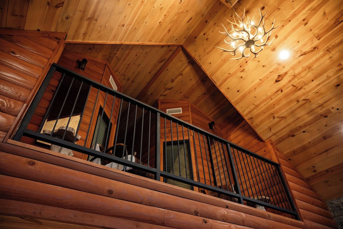 A wooden cabin interior with a balcony, a chandelier, and a window with blinds.