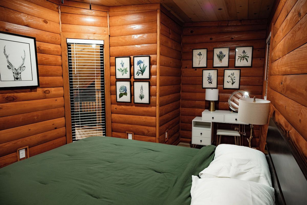 Ein Zimmer einer Holzhütte mit einem Bett, einem Nachttisch, einer Lampe und gerahmten Pflanzenbildern an der Wand.