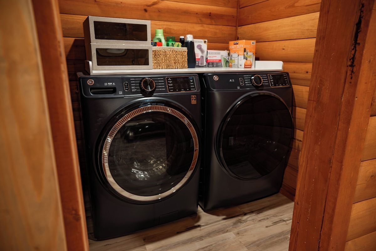 Un cuarto de lavandería con dos lavadoras de carga frontal, un microondas y varios productos de limpieza en un estante.
