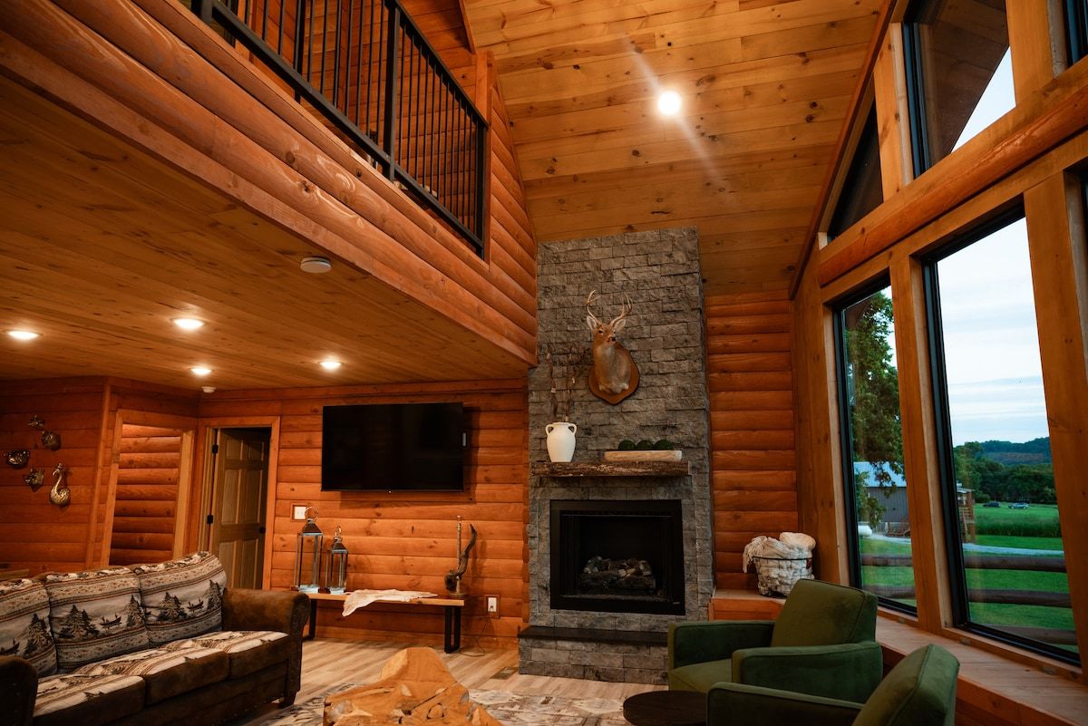 A log cabin interior with a stone fireplace, a mounted deer head, a television, and a large window offering a view of the outdoors.