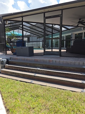 Un espacio exterior techado con escalones que dan a una piscina, rodeado de paredes de vidrio y un ventilador de techo.