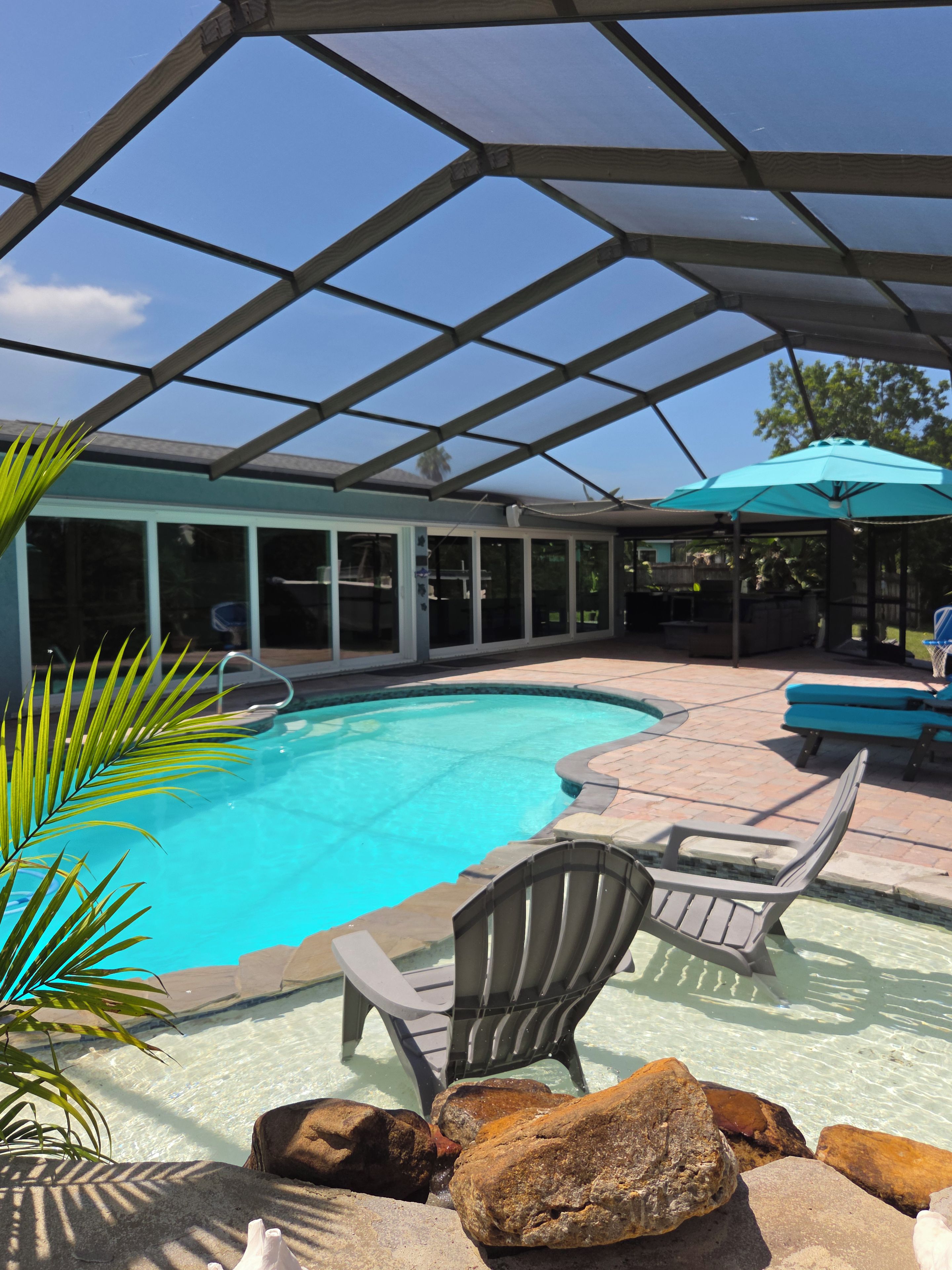 A swimming pool with a glass enclosure, lounge chairs, and a patio area.