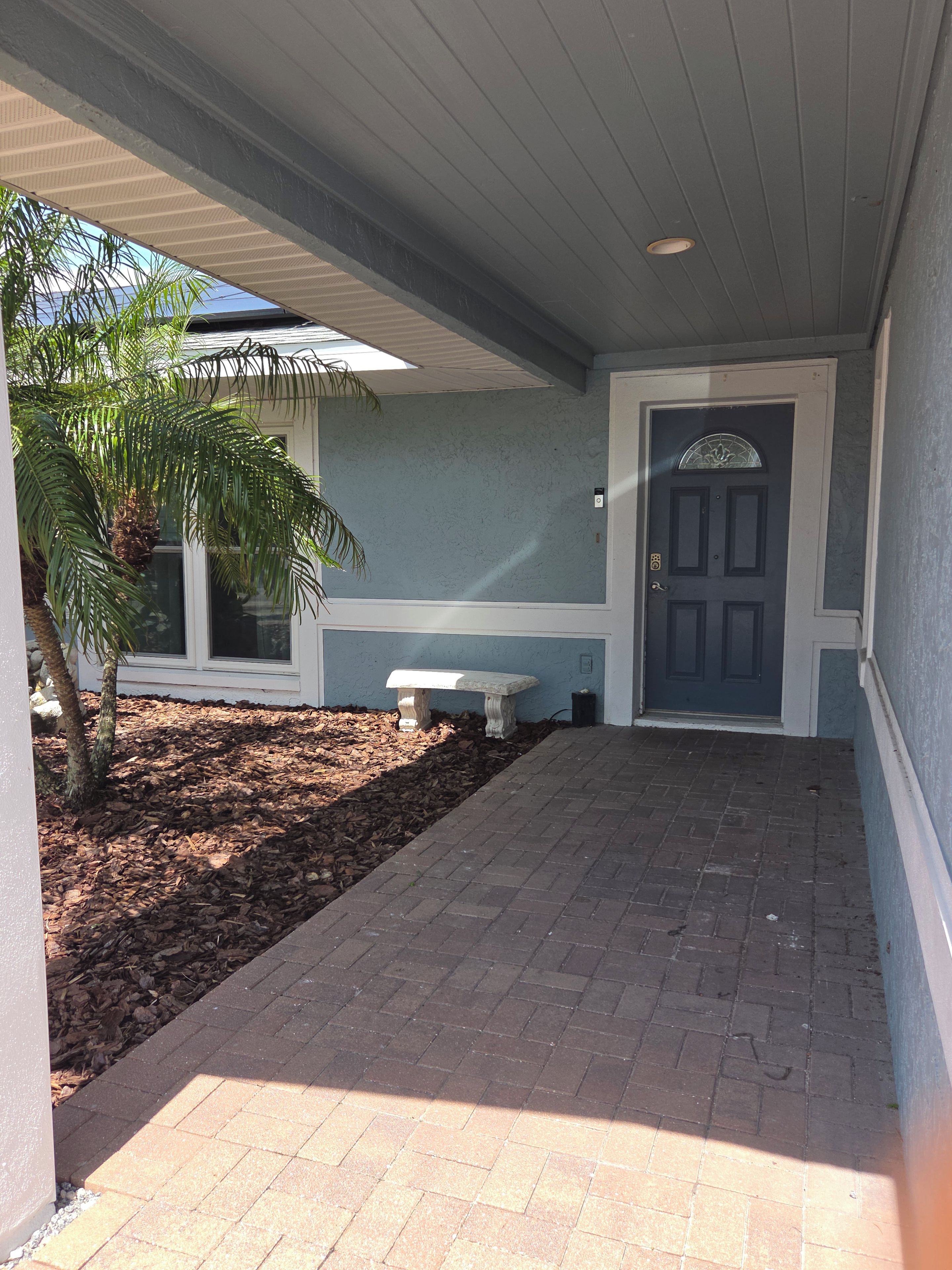 A covered porch with a bench, a door, and a palm tree.