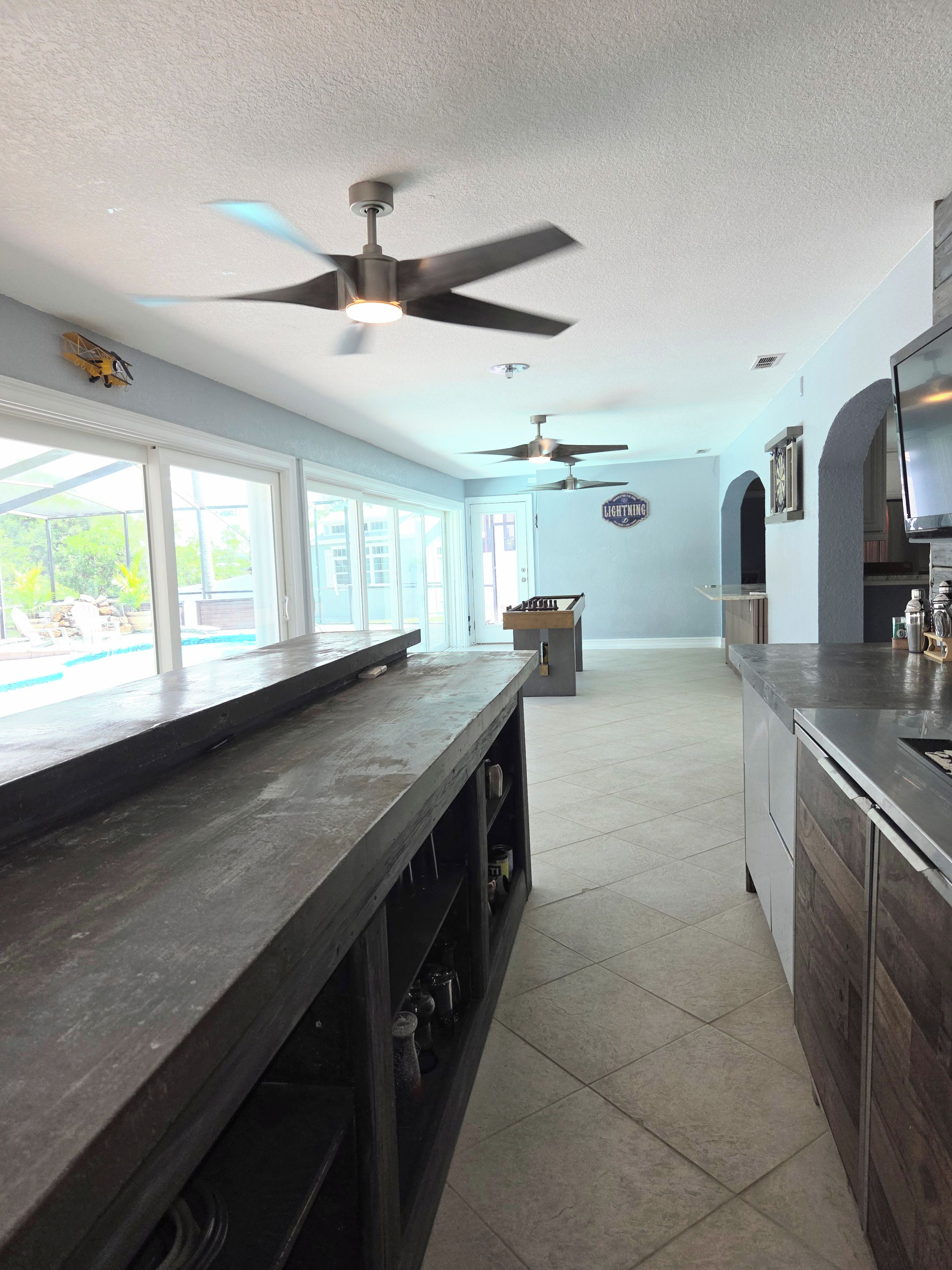 A spacious kitchen with a bar area, ceiling fans, and a view through large windows.