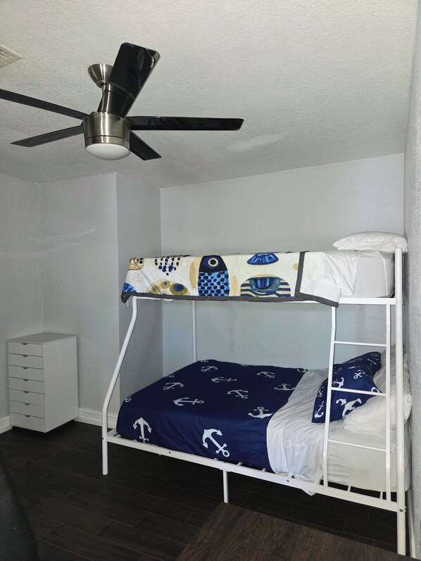Una cama litera con ventilador de techo, un aparador blanco y una colcha estampada.
