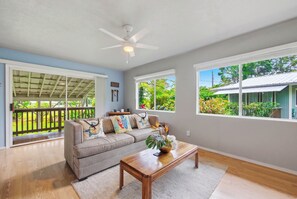 Ein Wohnzimmer mit einer Couch, einem Couchtisch und einem Deckenventilator. Es gibt Fenster mit Blick auf Grünflächen und ein Haus.