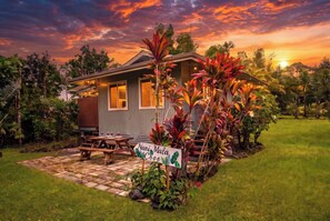 A house with a covered patio, a sign reading 'Nani Mala Cove', and a sunset in the background. - Island Rainforest Grove Two Cabin Combo (Keaau)