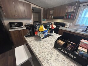 Una cocina con armarios de madera, cafetera, microondas y una barra de aperitivos con diversos alimentos.