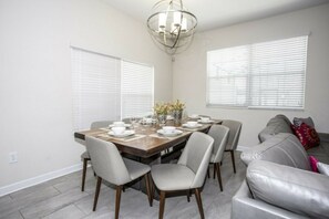 A dining area with a wooden table set for four, surrounded by chairs, and a sofa in the background. - 5BR Storey Lake | Pool, Mickey & Minions rooms (Kissimmee)
