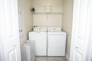 A laundry room with two white dryers and a washing machine, a basket on a shelf above, and a small covered area for drying clothes. - 5BR Storey Lake | Pool, Mickey & Minions rooms (Kissimmee)