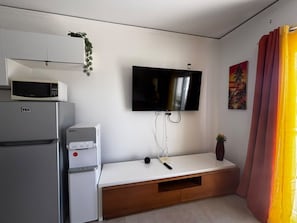A compact living space with a flat-screen TV mounted on the wall, a white cabinet with a microwave, a refrigerator, and a wooden bench with a small plant. - Sunset Vibes - Studio, pool views, resort access (Sosúa)