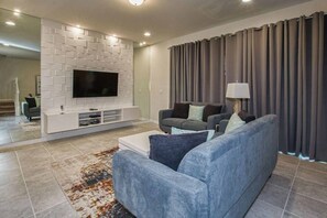 A modern living room with a flat-screen TV mounted on a white wall, a grey sofa with cushions, a coffee table, and a patterned rug. - 5BR Disney townhome | Pool & themed rooms (Kissimmee)