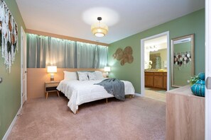 Une chambre à coucher comprenant un lit, des lampes de chevet, un luminaire au plafond, un miroir et une porte donnant sur une salle de bain.