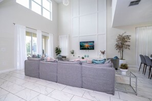 Ein geräumiges Wohnzimmer mit einem großen, grauen Ecksofa, einem flachbildfernseher an der Wand und einem Essbereich mit einem Glastisch und Stühlen.