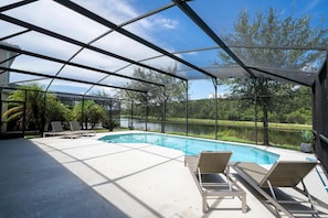 A covered outdoor swimming pool with lounge chairs, surrounded by a natural landscape and a body of water. - 6BR Sonoma Resort home | Pool & Cars game room (Kissimmee)