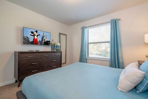 A bedroom with a bed, a dresser, a TV with a Mickey Mouse image, a window with curtains, and a lamp. - 6BR near Disney | Private pool, spa, movie room (Kissimmee)
