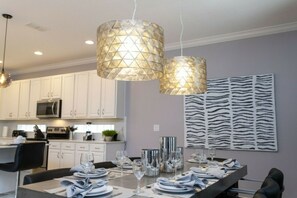 A modern kitchen with white cabinets, a dining area set for six with a long table, and pendant lights with a geometric pattern. - 6BR near Disney | Pool, spa, Mickey & Marvel (Kissimmee)