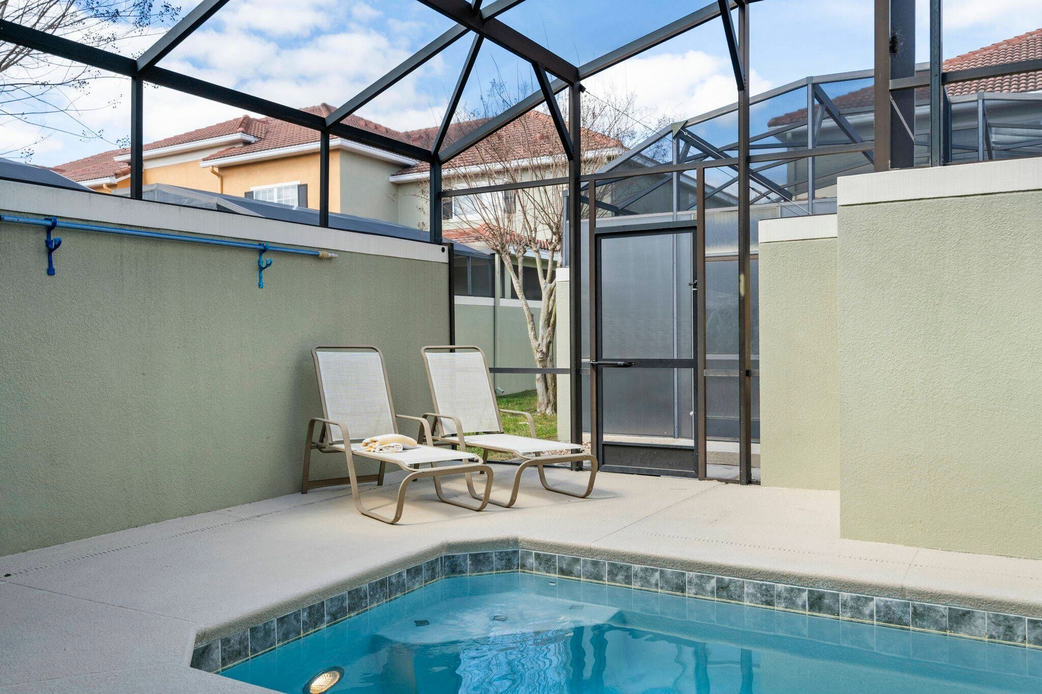 An outdoor swimming pool with two lounge chairs, a glass enclosure, and a view of neighboring houses.