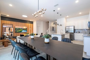Eine moderne Küche mit Edelstahlgeräten, ein Essbereich mit einem langen Tisch und Stühlen sowie ein Wohnbereich mit Fernseher und Holzverkleidung.