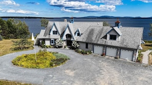 A two-story house with a steeply pitched roof, a garage, and a driveway leading to a landscaped area with a circular garden. - Peaceful Loft with Gazebo and Firepit (Surry)