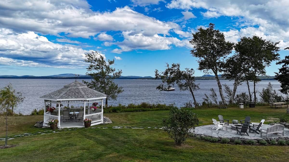 A gazebo with a patio, overlooking a lake with boats, trees, and distant mountains.