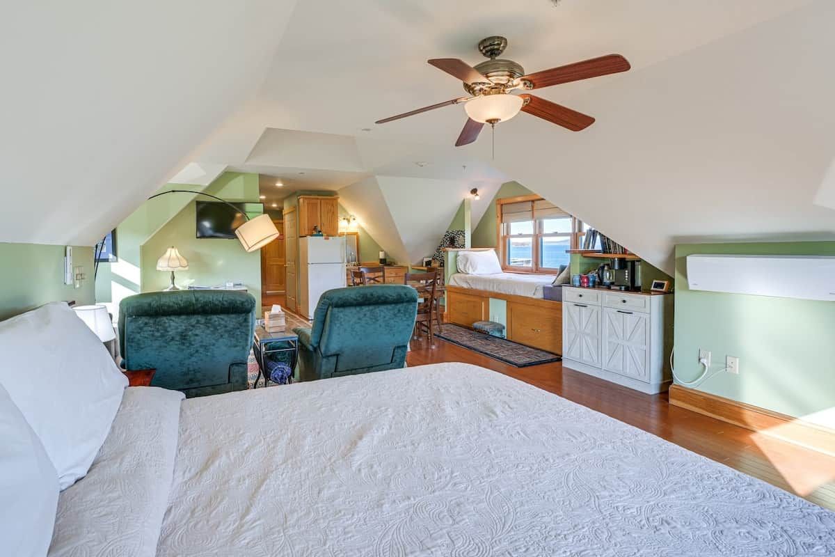 A bedroom with a bed, two chairs, a ceiling fan, and a kitchenette in the background.
